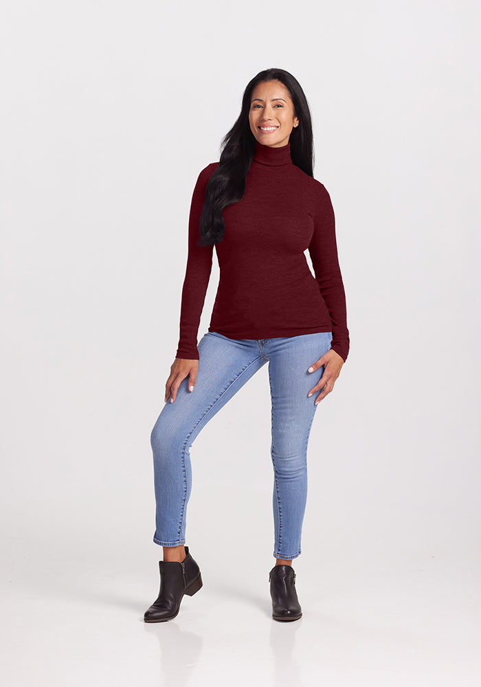 A woman with long dark hair is standing and smiling. She is wearing a burgundy Sage Turtleneck from Woolx, made from Australian Merino Wool, light blue jeans, and black ankle boots. She is posing with one leg slightly forward and one hand resting on her thigh against a plain white background.