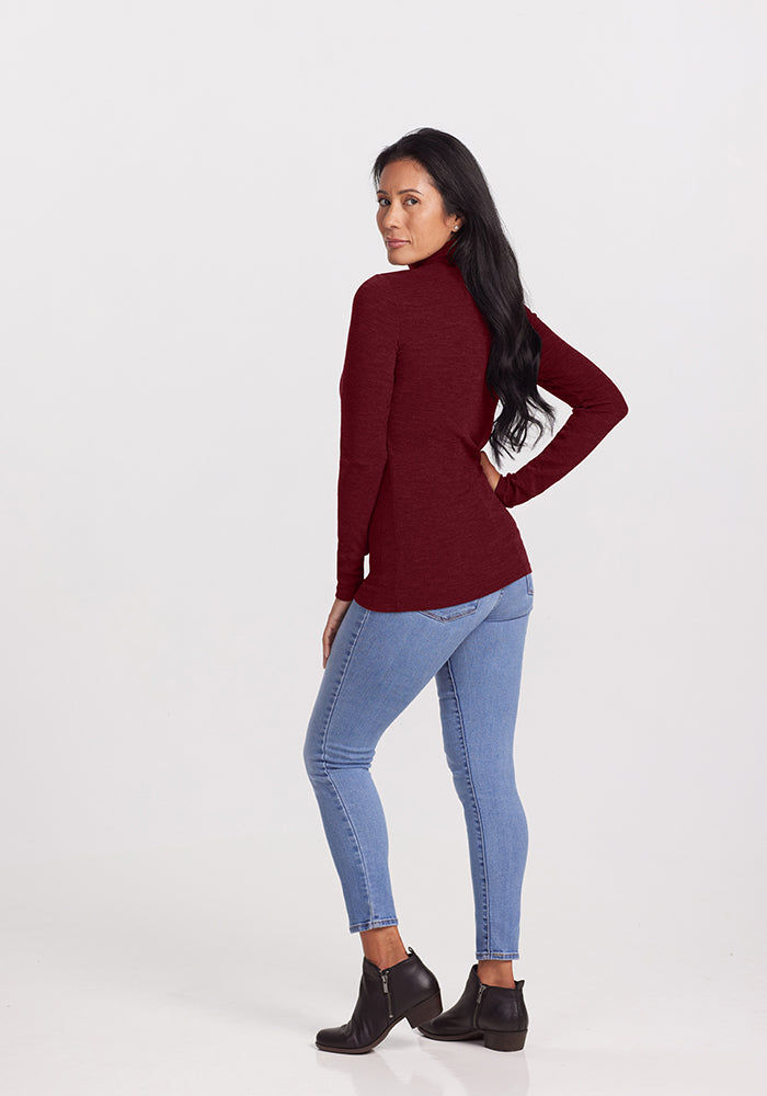 A woman with long dark hair, wearing a maroon Sage Turtleneck by Woolx, light blue jeans, and black ankle boots, is standing with her body facing to the side and her head turned towards the camera against a plain light background.