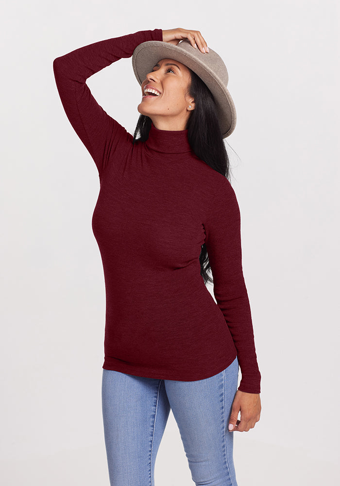 A woman wearing a maroon Woolx Sage Turtleneck and light blue jeans smiles and poses with one hand on her gray hat, the other resting by her side. Her long black hair is slightly tilted back, and the plain, light-colored background accentuates the outfit's moisture-wicking properties.