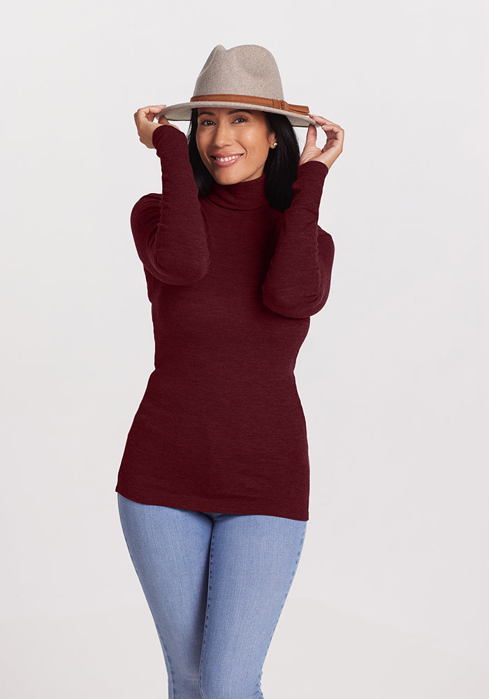 A woman wearing a maroon Woolx Sage Turtleneck made from Merino wool and light blue jeans smiles while holding a beige hat on her head with both hands. She stands against a plain white background.