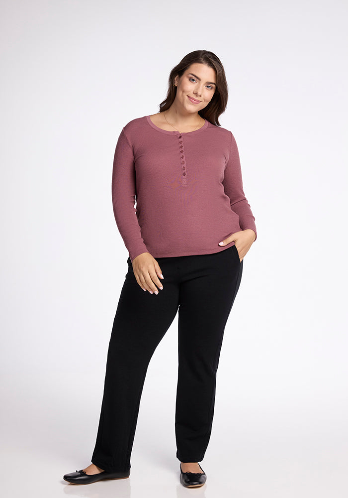 A woman with long, dark hair stands against a plain background. She is wearing a Reese Ribbed Top in mauve from Woolx, black pants, and black ballet flats. One hand is in her pocket while the other rests on her thigh, and she is smiling softly at the camera.