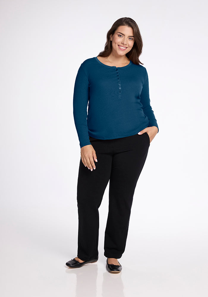 A woman with long dark hair smiles while standing. She is wearing a long-sleeve blue Reese Ribbed Top by Woolx and black pants. One hand rests in her pocket, and the other by her side. She is also wearing black shoes. The background is plain white.