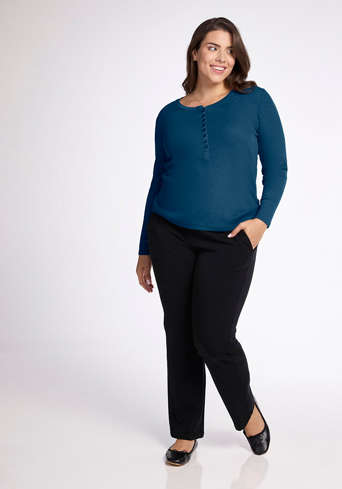 A woman with long brown hair is standing and smiling. She is wearing a blue Reese Ribbed Top from Woolx, made from Australian Merino Wool, paired with black pants. Black ballet flats complete her look against the plain white background.