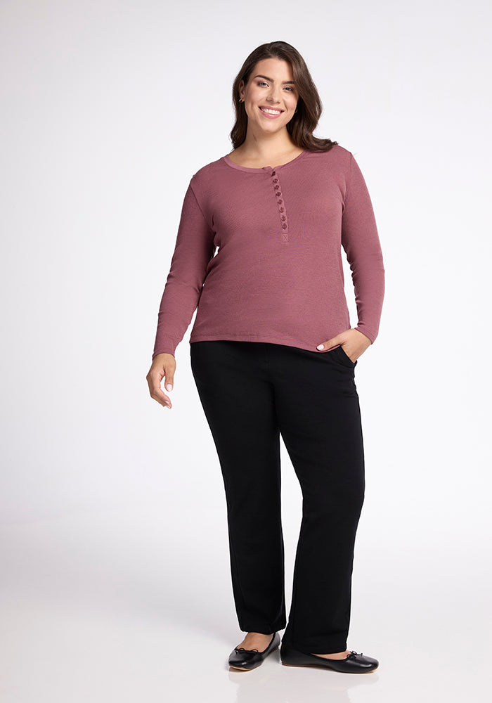 A woman stands facing the camera, smiling. She is wearing the Reese Ribbed Top by Woolx in maroon and black pants. She has one hand in her pocket and the other arm resting by her side. She is also wearing black flats. The background is plain and white.