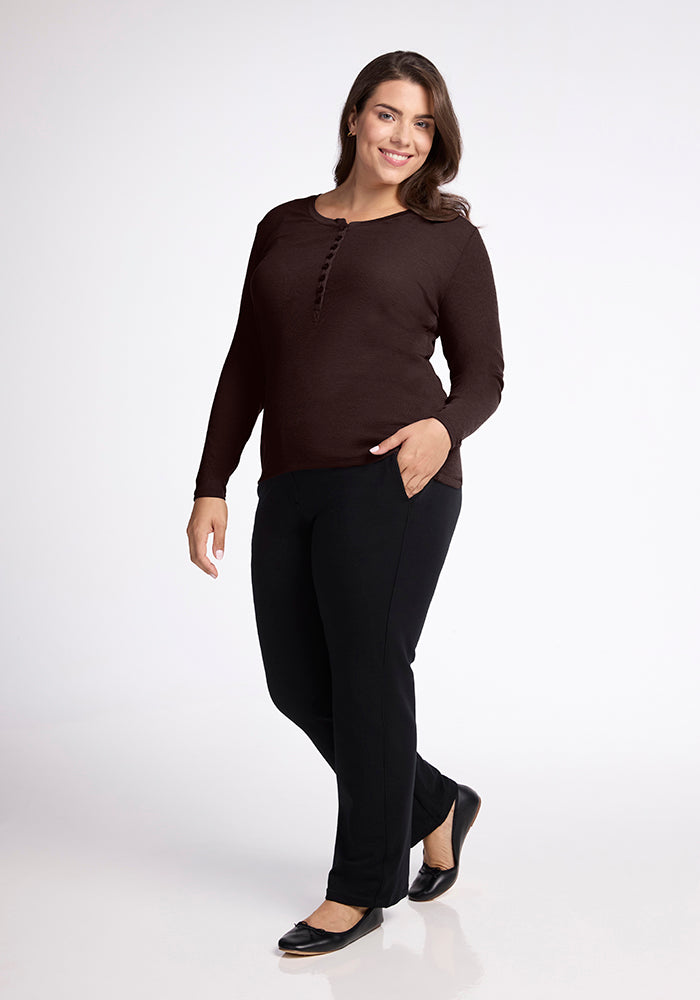 A woman with long, brown hair is smiling and standing with one hand in her pocket. She's wearing a Woolx Reese Ribbed Top made from Australian Merino Wool, paired with black pants and black flats. The background is plain white.