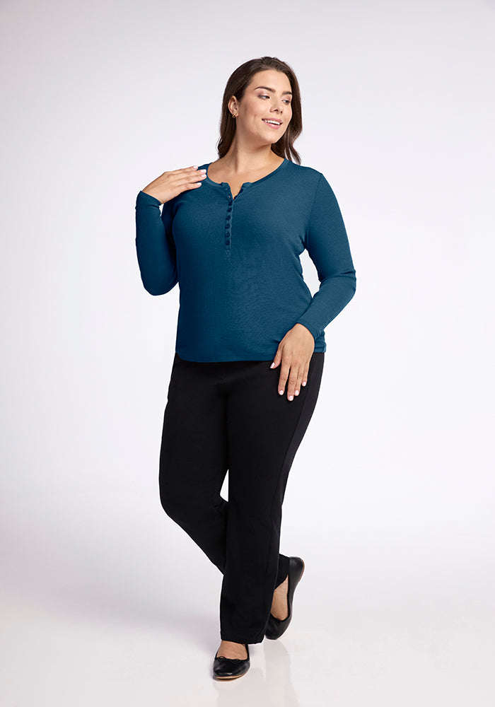 A woman stands and smiles with her left hand touching her shoulder, wearing a long-sleeve Reese Ribbed Top by Woolx made of Australian Merino Wool in teal, black pants, and black ballet flats. She has long dark hair and is standing against a plain white backdrop.