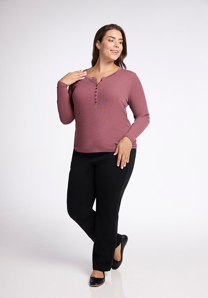 A woman with long dark hair is standing against a plain white background, wearing a Woolx Reese Ribbed Top and black pants. She is looking to the side and smiling, with one hand touching her neck and the other resting at her side.