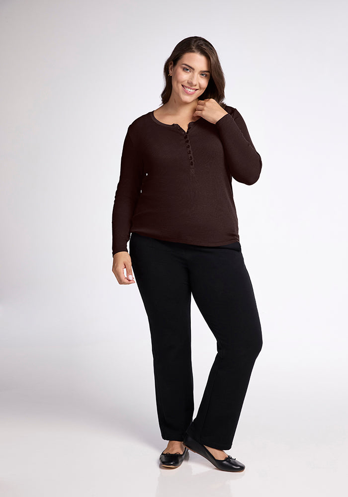 A woman with long dark hair stands smiling with her right hand touching her chin. She is wearing a long-sleeve brown Reese Ribbed Top from Woolx, black pants, and black flats. The background is a plain, light-colored studio setting.