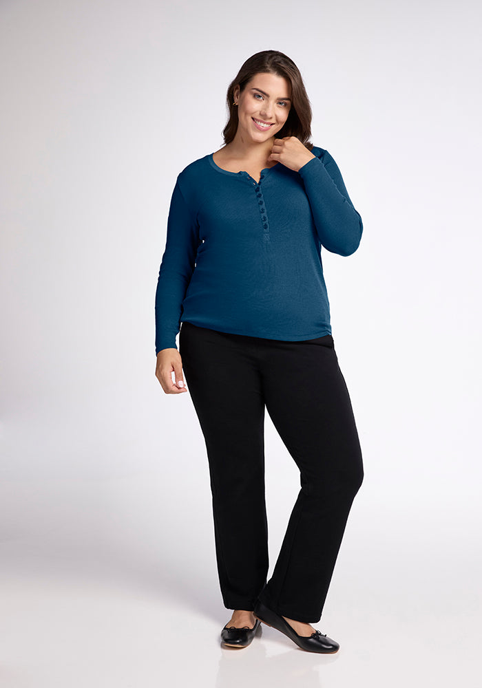 A woman stands against a plain background, smiling and looking at the camera. She is wearing a Reese Ribbed Top from Woolx, crafted from Australian Merino Wool in a long-sleeved, button-up henley style in teal. Complementing her top are black trousers and black shoes. One hand is touching her hair.