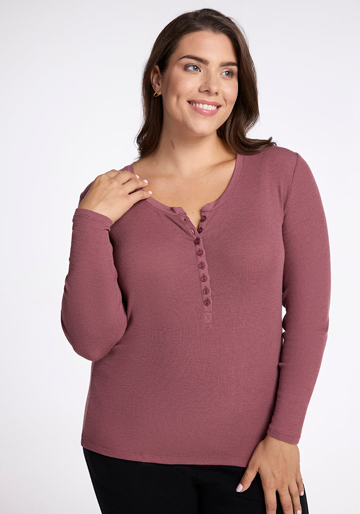 A woman with long brown hair smiles while wearing a rose-colored Reese Ribbed Top by Woolx. She is standing against a plain white background. 