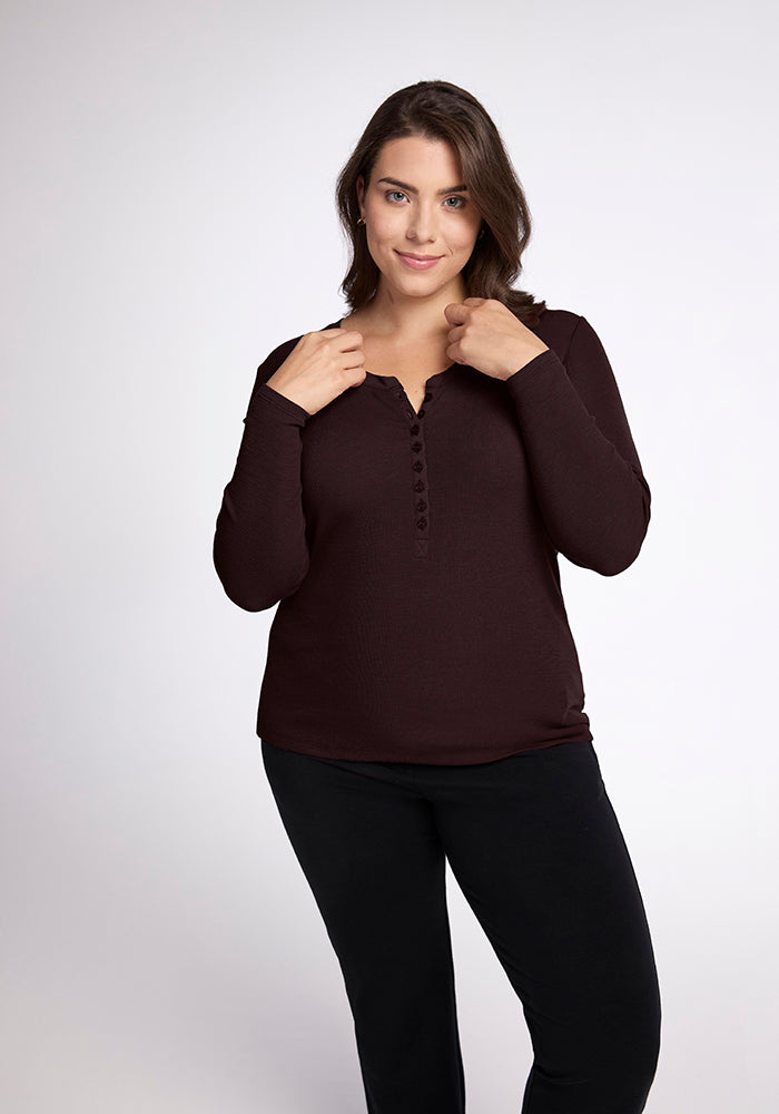 A woman with shoulder-length brown hair stands against a plain background, wearing a dark ribbed Reese Ribbed Top from Woolx made of Australian Merino Wool and black pants. She is holding the collar of her shirt with both hands and smiles slightly while looking at the camera.