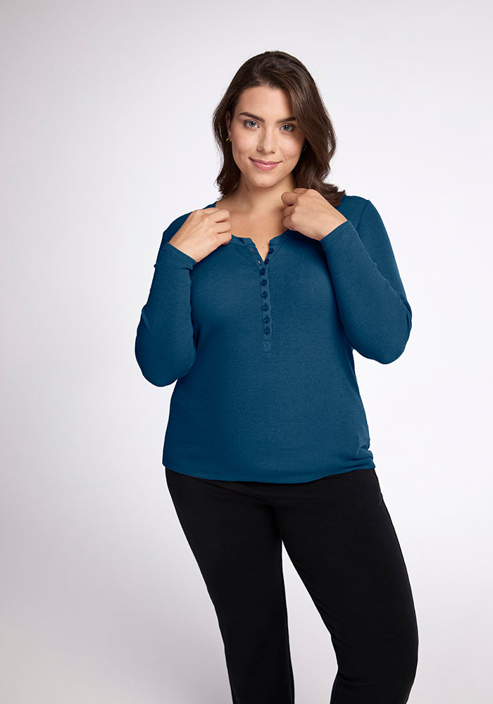 A woman with dark hair, wearing a long-sleeved, teal Reese Ribbed Top from Woolx made of Australian Merino Wool and black pants, stands against a plain background. She is gazing directly at the camera with a slight smile, holding the collar of her shirt with both hands. 