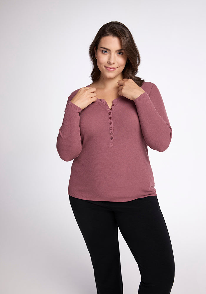 A woman with shoulder-length brown hair is wearing a Woolx Reese Ribbed Top in a rich maroon hue and black pants. She stands against a plain light gray background, facing the camera, her hands near the top buttons of her shirt, smiling slightly.