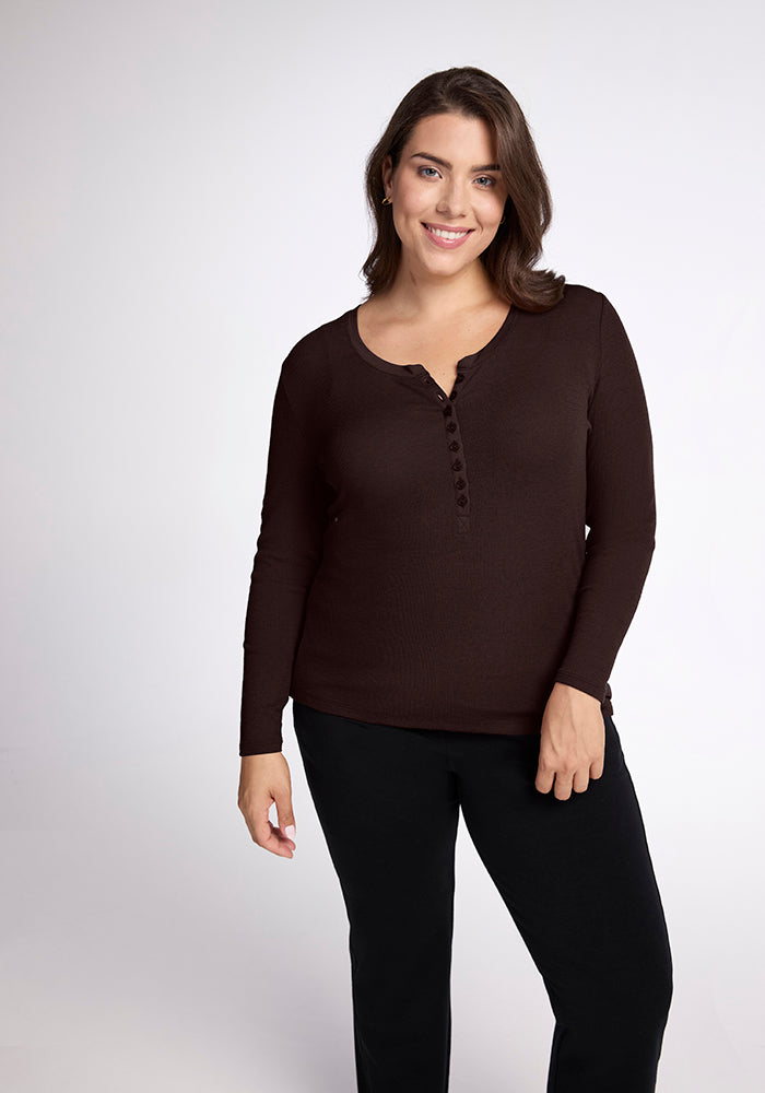 A person with shoulder-length brown hair is wearing a long-sleeved, dark brown Reese Ribbed Top by Woolx and black pants. They are standing against a plain white background, smiling, and looking towards the camera. 