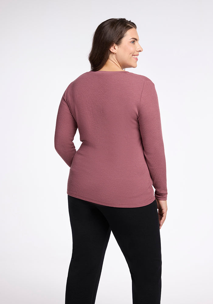 A person with long dark hair is standing and smiling, showing a partial side and back view. They are wearing a mauve-colored Reese Ribbed Top from Woolx and black pants. The background is plain white.