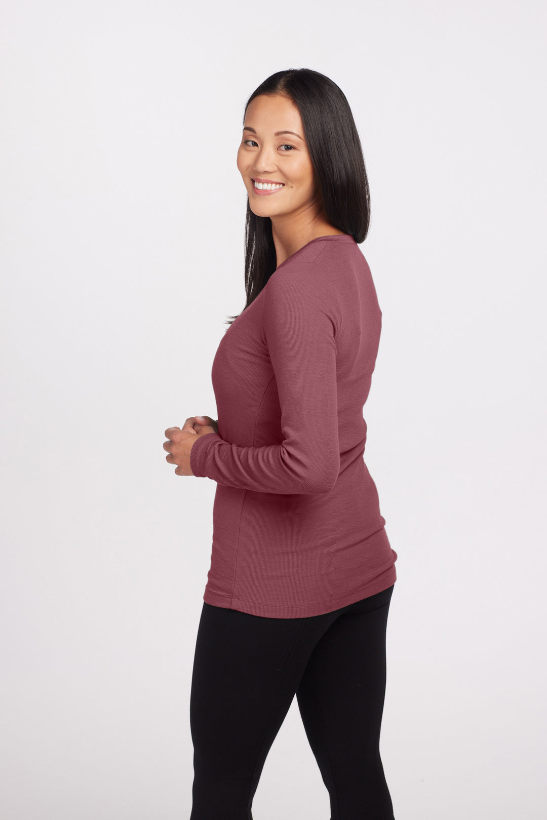 A woman with long black hair is standing sideways and smiling at the camera. She is wearing a maroon Reese Ribbed Top from Woolx, made of Australian Merino Wool, along with black pants. The background is plain white.