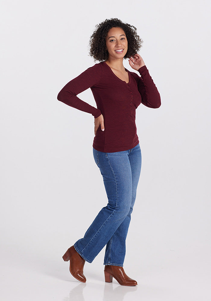 A person with curly hair smiles at the camera, wearing a cranberry melange Reese Ribbed Top by Woolx, along with blue jeans and brown heeled ankle boots. One hand is raised near their face while the other rests on their hip. They are standing on a plain white background.