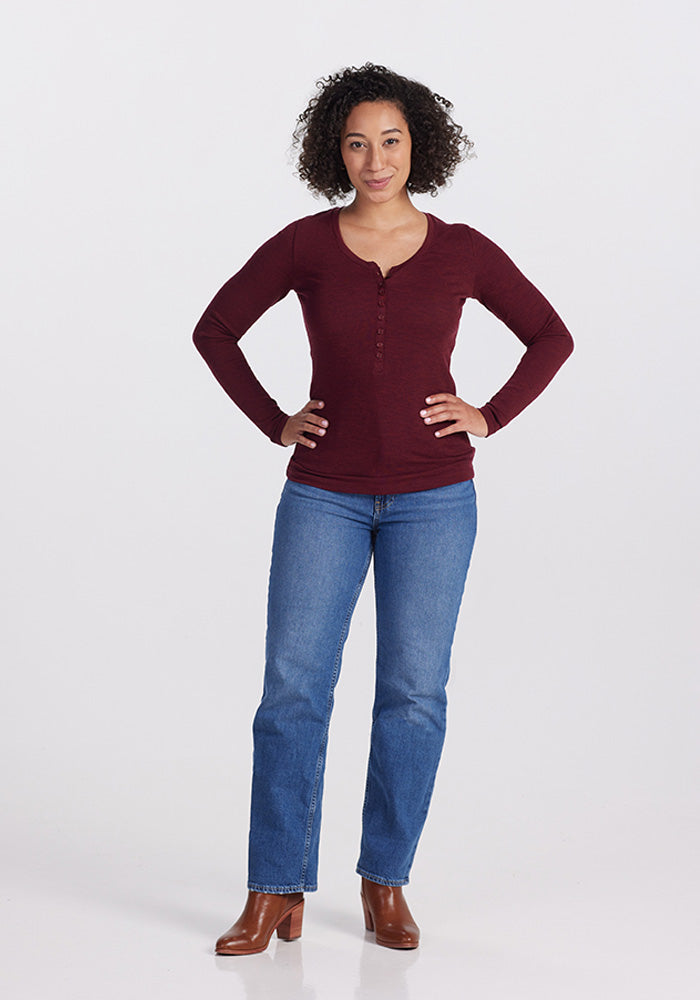 A person with curly hair stands confidently with hands on hips, wearing a Reese Ribbed Top - Cranberry Melange from Woolx, blue jeans, and brown ankle boots. The background is plain white.