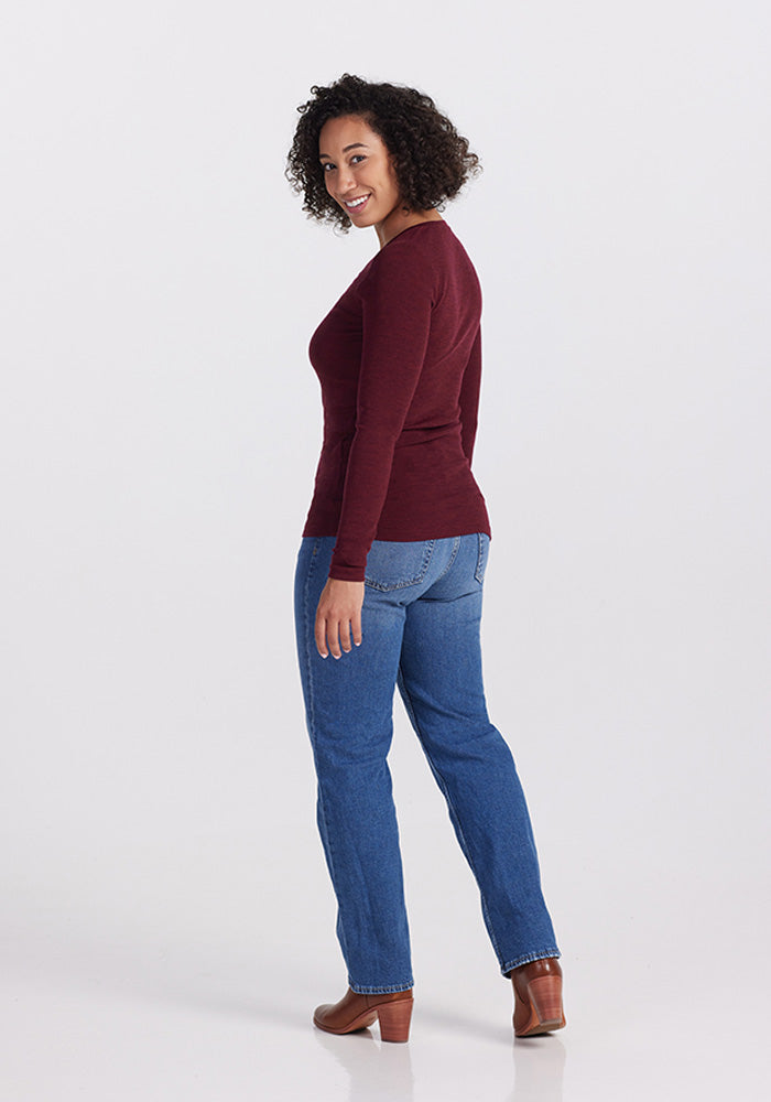 A woman with curly hair is standing and smiling at the camera. She is wearing a long-sleeved Cranberry Melange Reese Ribbed Top by Woolx, blue jeans, and brown boots. The background is plain white.