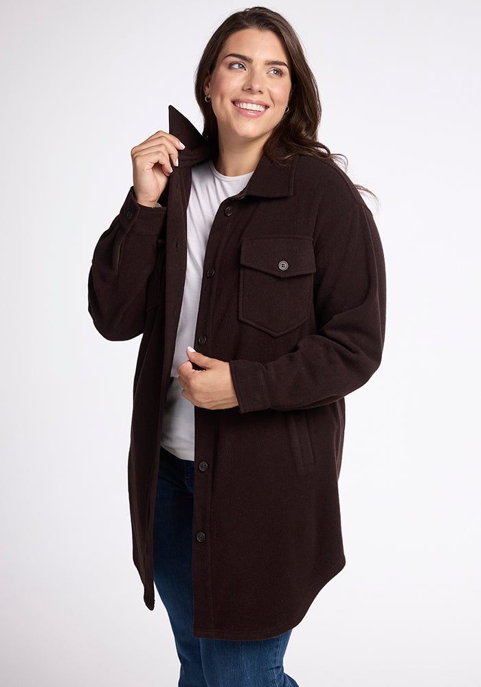 A person with long dark hair is smiling and looking to the side while wearing the Sutton Shacket from Woolx, a dark brown button-up coat made from Merino wool. The coat features large front pockets, and the person is casually holding onto its collar and front. They are also wearing blue jeans and a white shirt. 