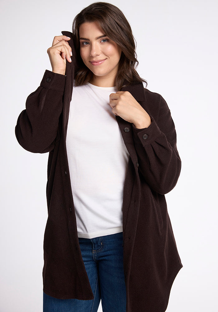 A woman with long brown hair smiles softly while adjusting the collar of her dark brown button-up shirt. She is wearing a white T-shirt underneath and blue jeans, standing against a plain white background where the Sutton Shacket by Woolx hangs to the side.