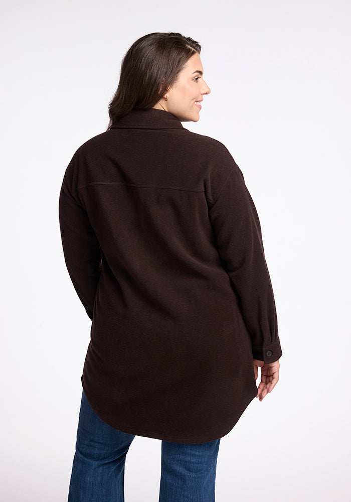 A woman with long dark hair is standing and facing away from the camera. She is wearing a dark brown Sutton Shacket by Woolx and blue jeans. The background is plain white.