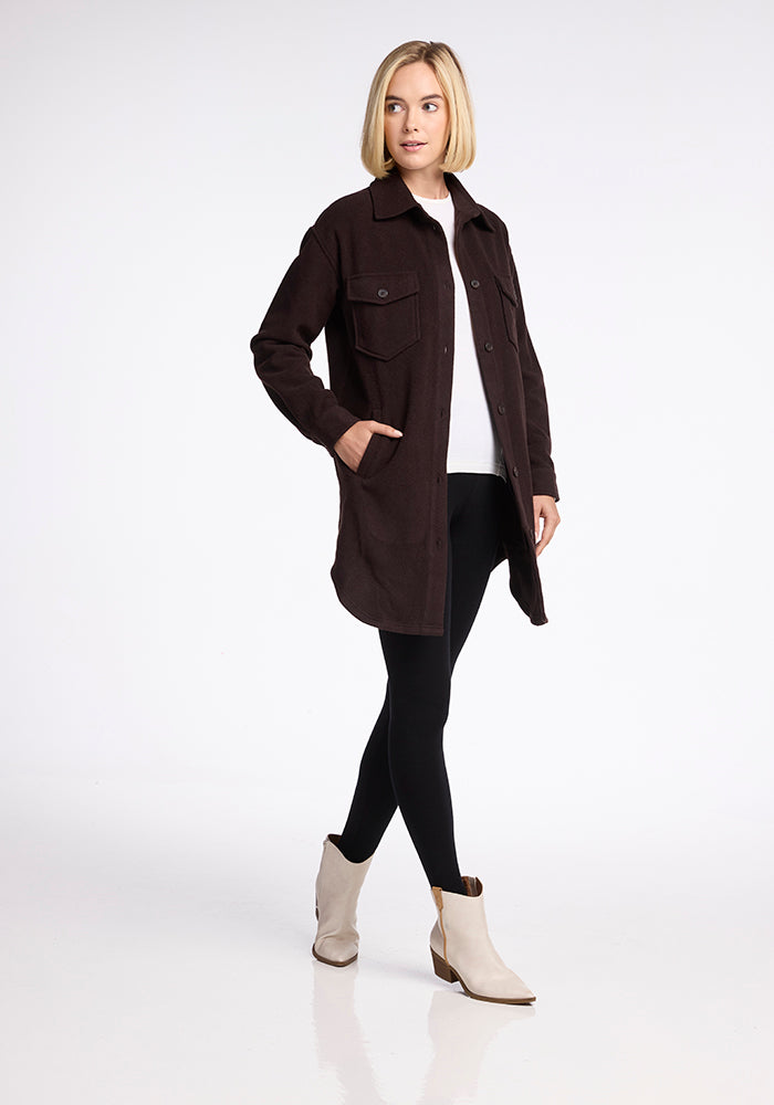 A woman with shoulder-length blonde hair is wearing a long, dark brown button-up Woolx Sutton Shacket with two chest pockets, black leggings, and light-colored ankle boots. She is standing against a plain white background with her hands in her jacket pockets, looking to the side.