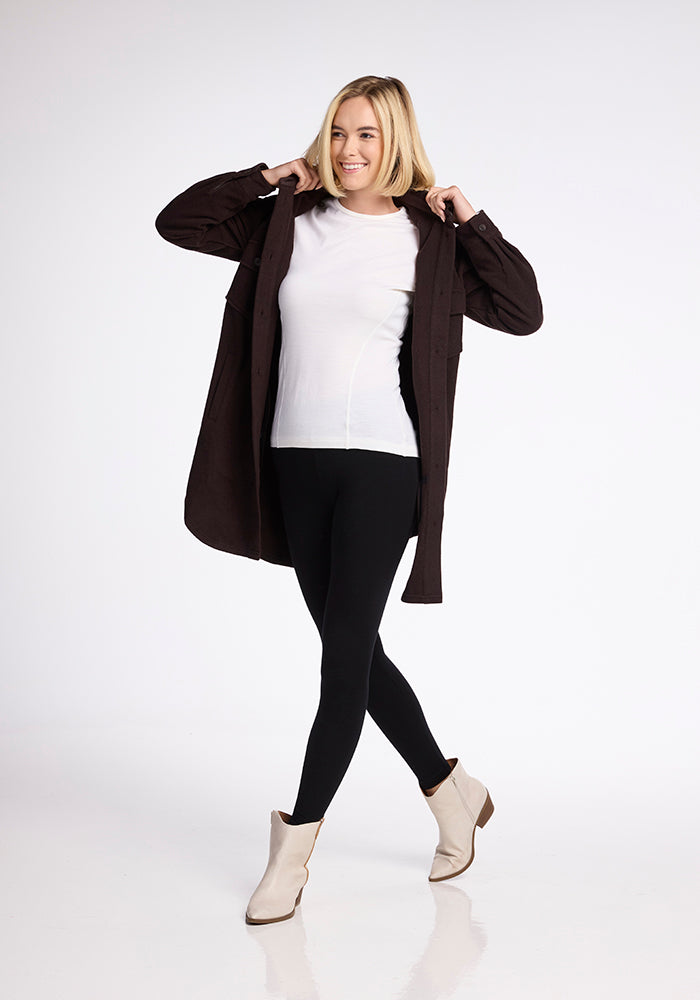 A person with blonde hair is smiling and looking off to the side while posing. They are wearing the Sutton Shacket by Woolx over a merino wool white shirt, black leggings, and beige ankle boots.