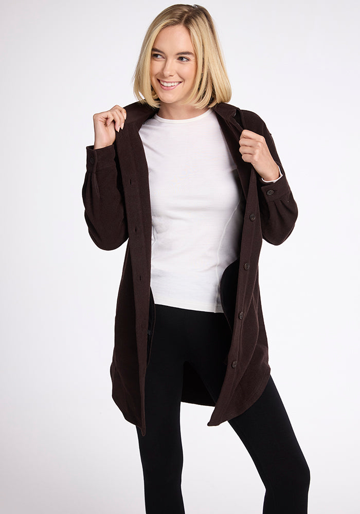 A person with shoulder-length blonde hair is smiling while wearing a dark brown Sutton Shacket by Woolx over a white shirt and black pants. 