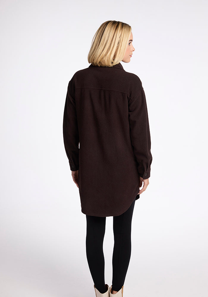 A person with short blonde hair is standing with their back to the camera, wearing a long, dark brown Woolx Sutton Shacket and black leggings. The background is a plain white studio backdrop.