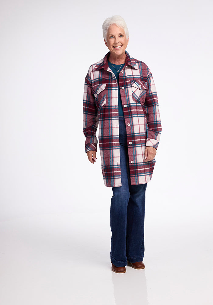 An older adult with short white hair is wearing a dark green top, blue jeans, and brown shoes. They are smiling and standing against a plain white background, showcasing their Woolx Sutton Shacket with its distinctive red and white plaid pattern.