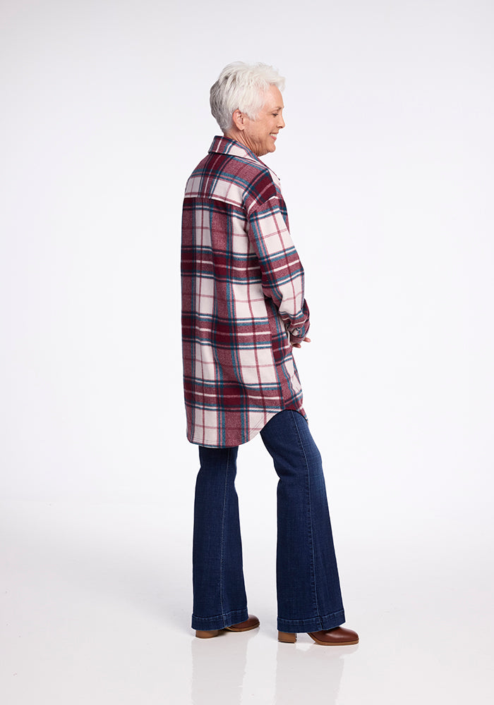 An older person with short white hair stands facing away, smiling and looking to the side. They are wearing a Woolx Sutton Shacket, which features a red, white, and blue plaid design in Merino wool. The shacket is paired with dark blue jeans and brown shoes. The background is plain white.