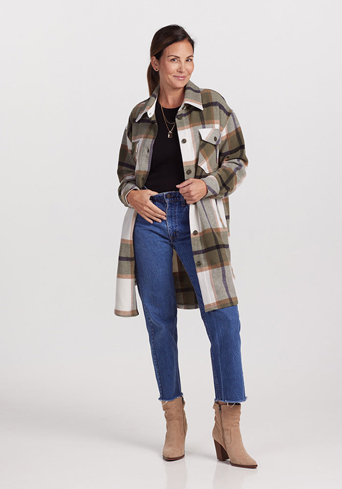 A person stands against a plain background wearing the Woolx Sutton Shacket in Forest Plaid, a black top, blue jeans, and tan ankle boots. They are smiling, with one hand in their pocket and the other resting on their hip.