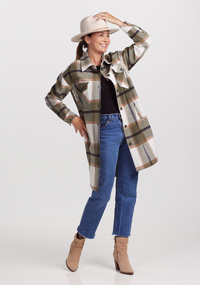 A woman stands smiling, donning a beige hat and the Sutton Shacket in Forest Plaid by Woolx over a black top, blue jeans, and beige ankle boots. She holds her hat with one hand and places the other on her hip against a plain white background.