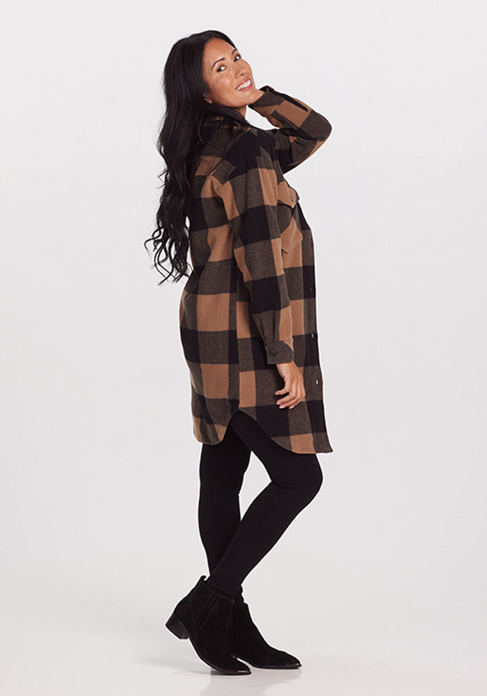 A person with long dark hair is smiling and posing with one hand on their hip and the other behind their head. They are wearing the Sutton Shacket - Camel Checkered by Woolx, black leggings, and black ankle boots. The background is plain white.