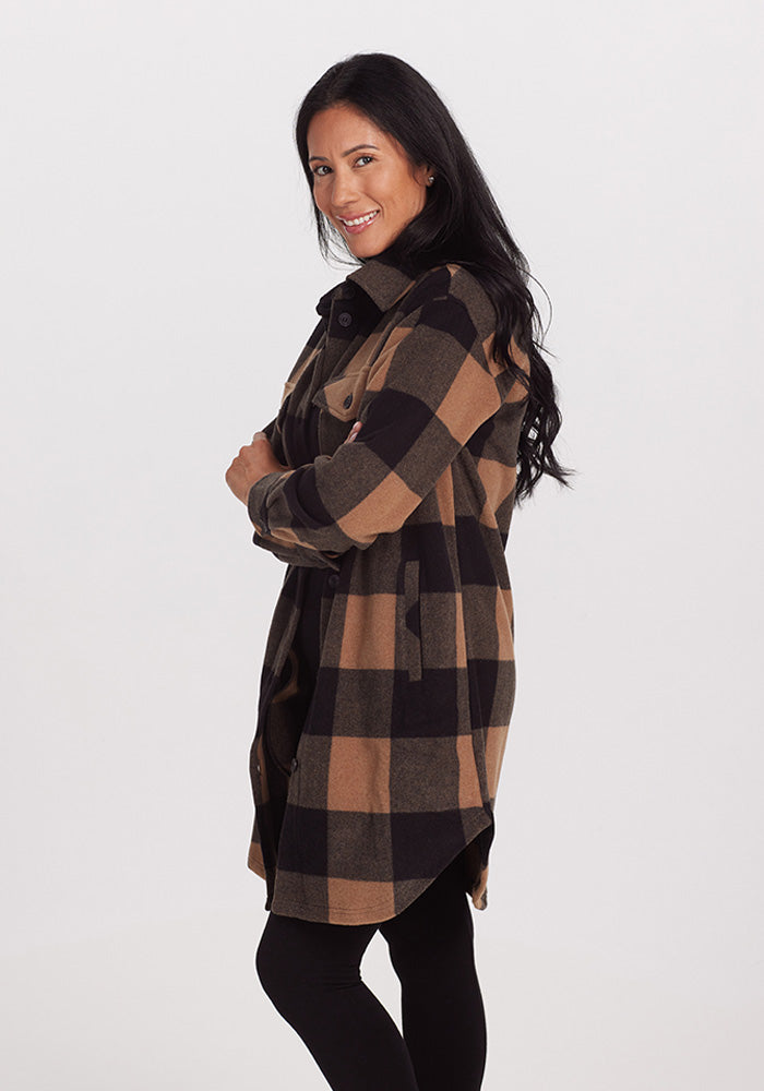 A woman with long dark hair is smiling and posing in profile view. She is wearing a Sutton Shacket in Camel Checkered, from the brand Woolx, along with black leggings, with her arms crossed casually. The background is plain white.