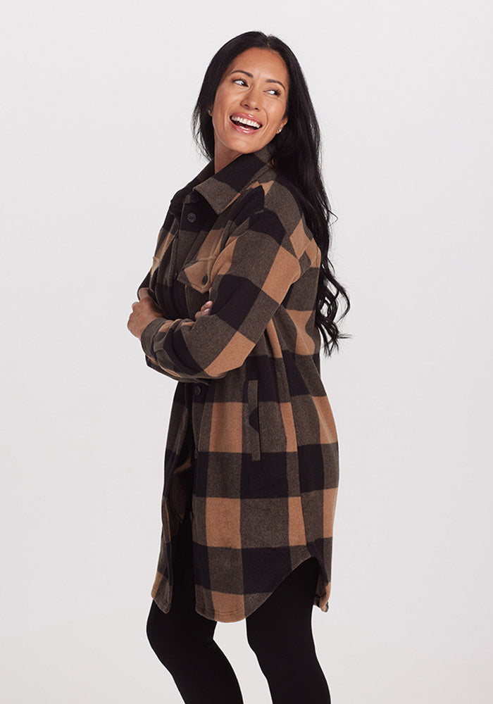 A woman with long black hair is smiling and looking off to the side. She is wearing the Woolx Sutton Shacket in Camel Checkered over black leggings. The background is plain white.