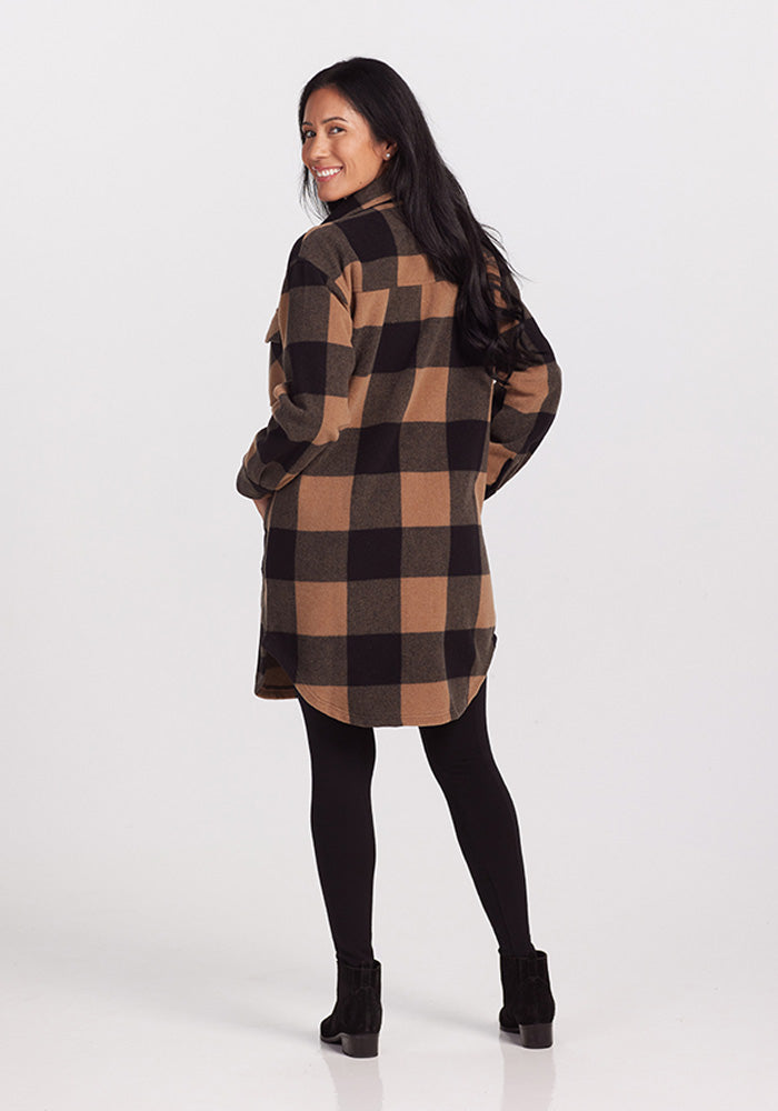 A person with long hair smiles while wearing the Sutton Shacket in Camel Checkered by Woolx over black leggings and ankle boots. They are standing on a plain, light background, looking back over their shoulder.