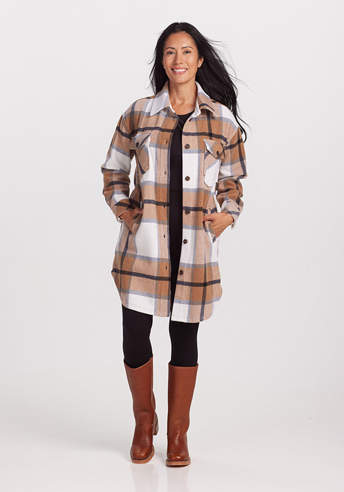 A person with long, dark hair is smiling while wearing the Woolx Sutton Shacket in Camel Plaid, featuring a pattern of brown, white, and black checks. They are also dressed in black pants and brown knee-high boots. The ultra-heavyweight merino wool of the shacket adds warmth to its stylish plaid design. The background is plain white.