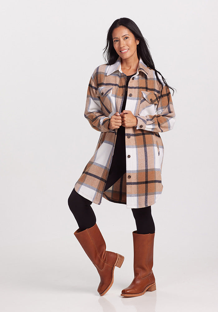 A woman with long dark hair stands smiling against a plain background. She is wearing the Woolx Sutton Shacket in Camel Plaid, black leggings, and brown knee-high boots. She holds the shacket's lapels with both hands.