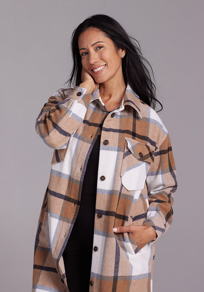 A woman with long dark hair smiles while posing, one hand touching her face and the other in the pocket of her long, Camel Plaid Sutton Shacket from Woolx. The shacket features a brown, white, and gray checkered pattern. The background is plain and light gray.