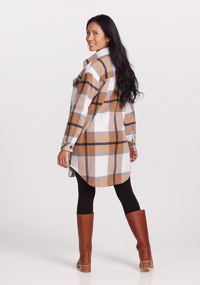 A woman with long dark hair is standing and smiling, wearing the stylish Sutton Shacket - Camel Plaid by Woolx. She pairs this fashionable winter wear with black leggings and knee-high brown boots. The background is plain white, and she is seen from a slight side angle, looking back.