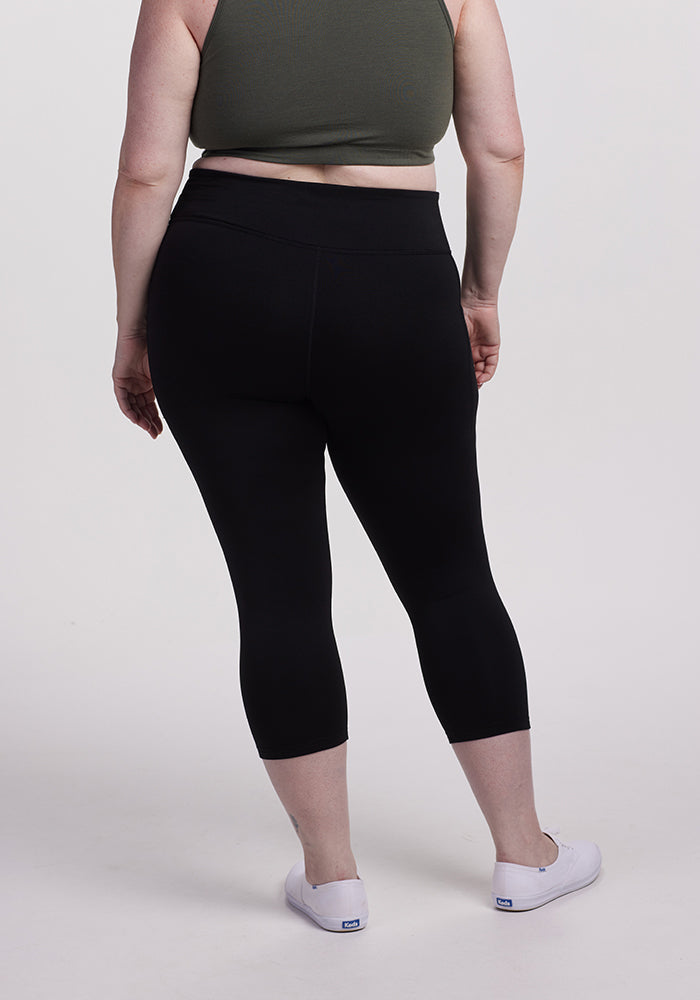 A person is standing with their back to the camera, embodying the athleisure style in a dark green crop top and the McKenna Capris - Black by Woolx. They sport white sneakers against a plain, light-colored background.