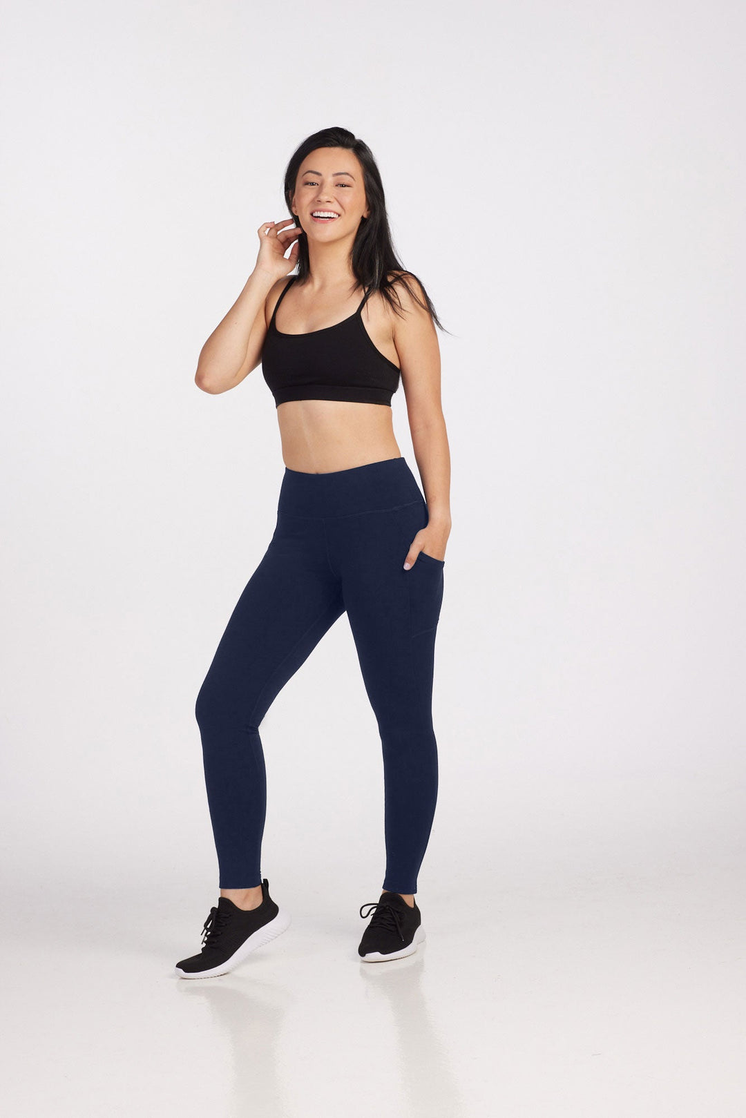 A woman stands smiling against a plain background. She has long dark hair and is dressed in workout attire, including a black sports bra and navy blue Piper Pocket Leggings made from Australian Merino Wool by Woolx. She has one hand on her hip and the other touching her ear.