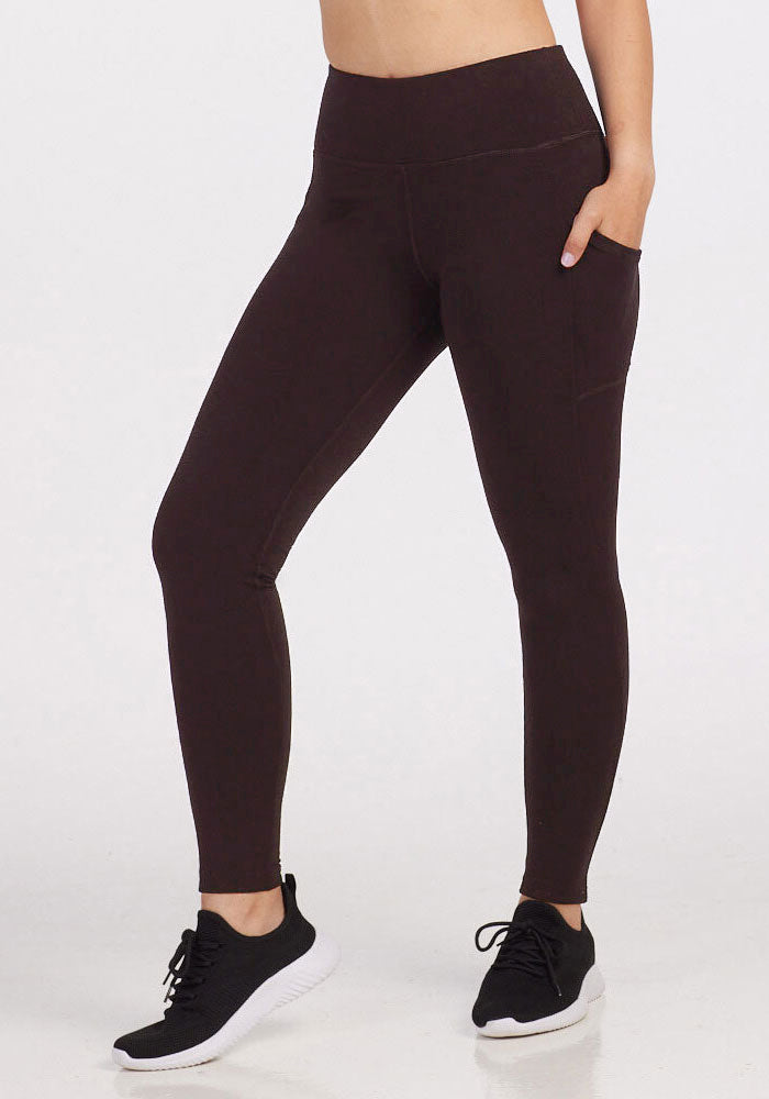 A person wearing Woolx's Piper Pocket Leggings in high-waisted, dark brown style with hands in the side pockets, paired with black athletic shoes on a white background. The upper part of the body is not visible. 