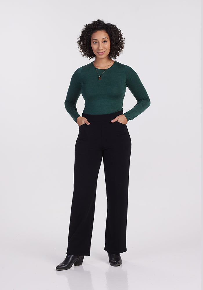 A petite woman with curly hair stands confidently, wearing a dark green long-sleeve top and Ellie Wide Leg Pants - Petite from Woolx in black Merino wool, paired with black boots. Her hands are in her pockets as she smiles against the plain white background.