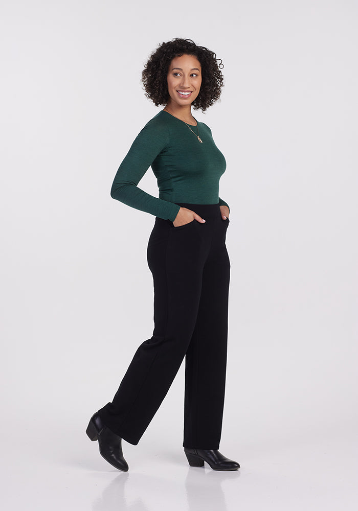 A woman with curly hair is smiling while standing sideways. She wears a green long-sleeve top, Ellie Wide Leg Pants Petite in black from Woolx, and black shoes, with her hands in her pockets. The background is plain white.