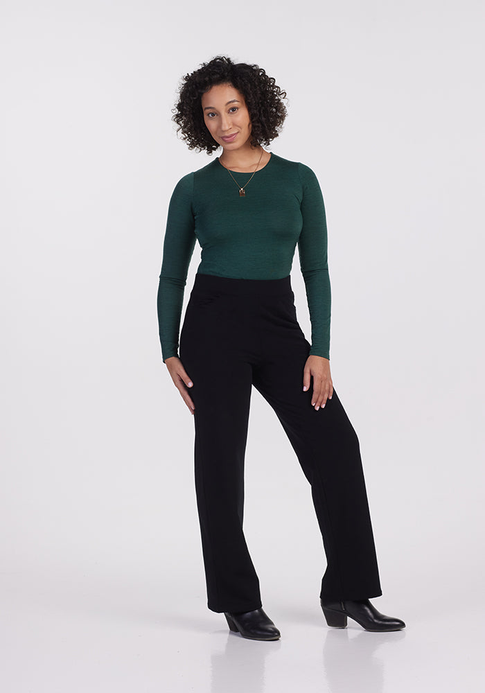 A person with curly hair stands against a white backdrop wearing a dark green Merino wool long-sleeve top, Ellie Wide Leg Pants in black by Woolx, and black boots. They have one hand on their hip and are accessorized with a gold necklace, exuding style in their Woolmark® Certified outfit.