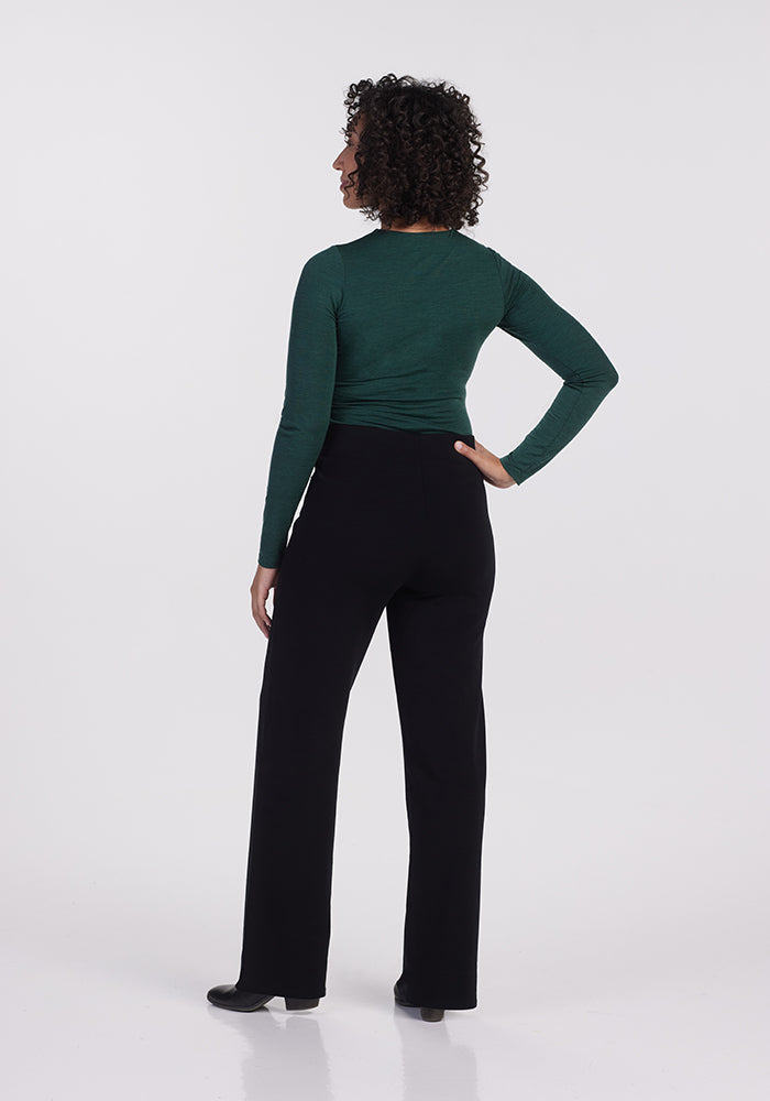 A person with curly hair stands facing away, wearing a long-sleeved green top and the Ellie Wide Leg Pants Petite in black from Woolx. The background is plain white.