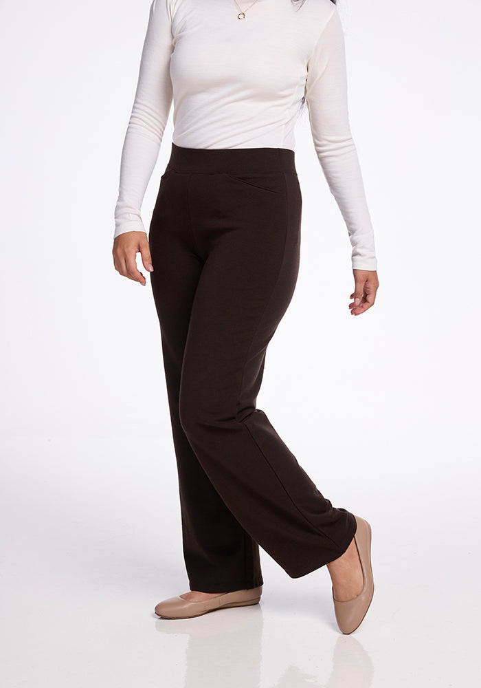 A person wearing a white merino wool long-sleeve top and Ellie Wide Leg Pants in French Roast by Woolx stands with one foot slightly raised. They are also wearing beige flats. The background is a plain white studio setting. 